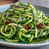 Vibrant spiralized zucchini mixed with homemade pesto and sweet cherry tomatoes, served with a sprinkle of Parmesan and fresh basil.  