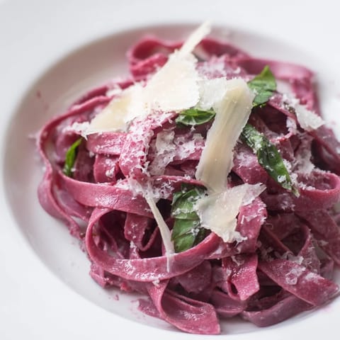 A close-up of homemade beet pasta noodles, garnished with fresh herbs and grated Parmesan, served warm on a rustic plate.  
